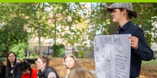 Draußen. Bäume mit grünen Blättern im Hintergrund. Ein junger Mann mit Hut hält ein Plakat. Um ihn herum sitzen mehrere junge Frauen.