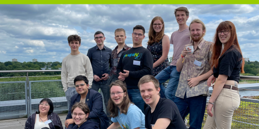 Eine Gruppe junger Menschen steht draußen an einem Stahlgeländer. Alle lächeln in die Kamera.