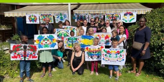 Kinder und Erwachsene stehen in einer Gruppe auf dem Rasen und halten selbstgemalte Bilder in die Kamera